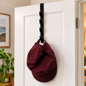 May include: A black over-the-door organizer rack featuring five integrated hooks arranged vertically. A solid dark maroon baseball cap is hanging from the bottom hook of the rack, which is mounted securely over the top edge of a white interior door.