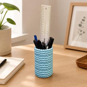 Puede incluir: Un portalápices azul claro con una textura similar a una cuerda, que contiene bolígrafos negros, un bolígrafo azul y una regla transparente. La regla está marcada con pulgadas y centímetros. El portalápices es cilíndrico y está sobre un escritorio de madera.