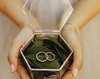 May include: A hexagonal glass ring box with a hinged lid, suspended by a chain. The box is filled with green leaves and two gold wedding bands.