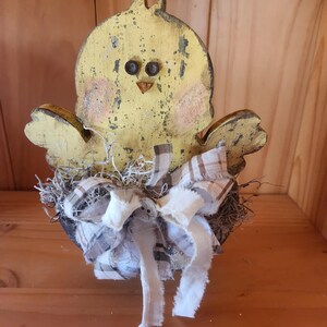 May include: A yellow wooden chick with black eyes and a brown beak sits in a nest made of brown twigs and white and brown fabric.