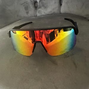 May include: Black-framed sunglasses with a vibrant, rainbow-colored lens. The lenses transition from red to yellow to blue, offering a bold and stylish look. The sunglasses have a sporty, wraparound design.