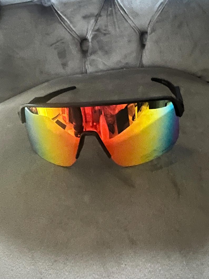 May include: Black-framed sunglasses with a vibrant, rainbow-colored lens. The lenses transition from red to yellow to blue, offering a bold and stylish look. The sunglasses have a sporty, wraparound design.
