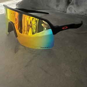 May include: Black sports sunglasses with a mirrored, multi-colored lens. The lenses transition from yellow to blue. The frame is black with a red logo on the temple. The text "PRIZM" is visible on the lens.
