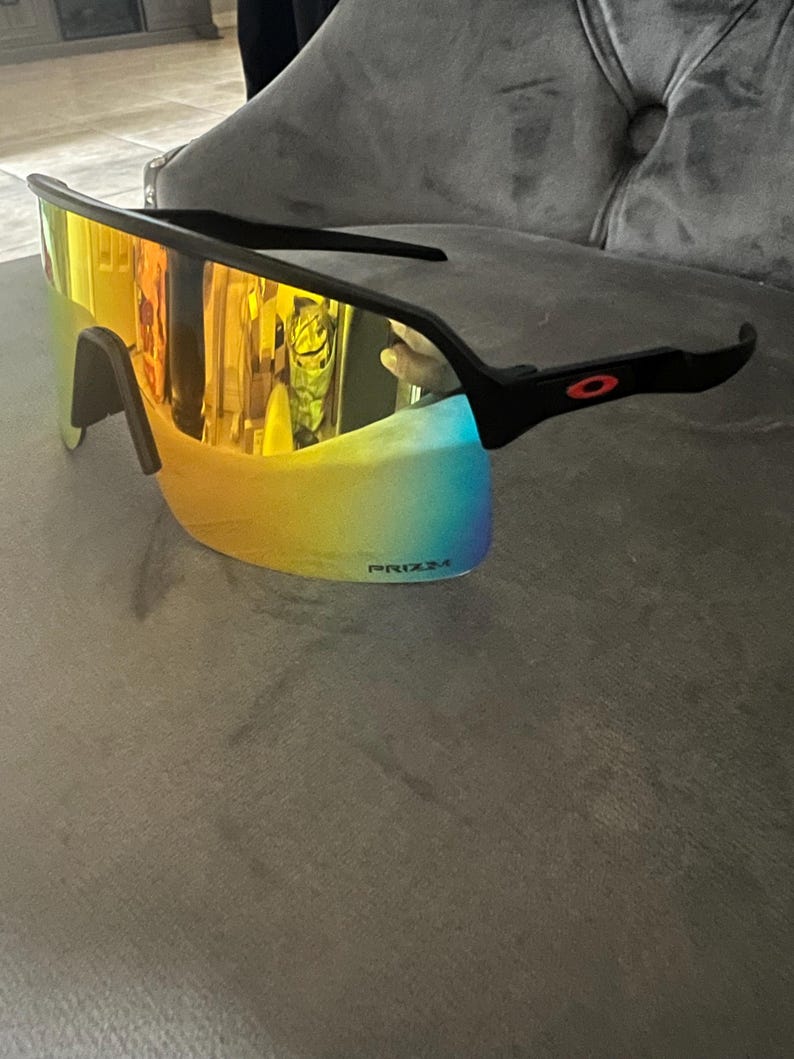 May include: Black sports sunglasses with a mirrored, multi-colored lens. The lenses transition from yellow to blue. The frame is black with a red logo on the temple. The text "PRIZM" is visible on the lens.
