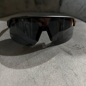 May include: Black sport sunglasses with a sleek, modern design. The sunglasses have a single-lens style with a dark tint and a black frame. The bridge of the sunglasses is a V-shape. The sunglasses are resting on a gray surface.