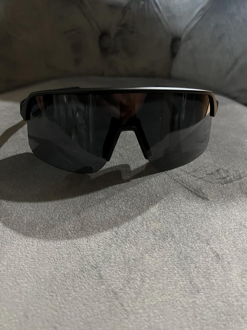 May include: Black sport sunglasses with a sleek, modern design. The sunglasses have a single-lens style with a dark tint and a black frame. The bridge of the sunglasses is a V-shape. The sunglasses are resting on a gray surface.