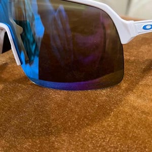 May include: White-framed sunglasses with a wraparound design. The lenses have a mirrored finish with blue and purple hues. The sunglasses are set against a brown textured surface.