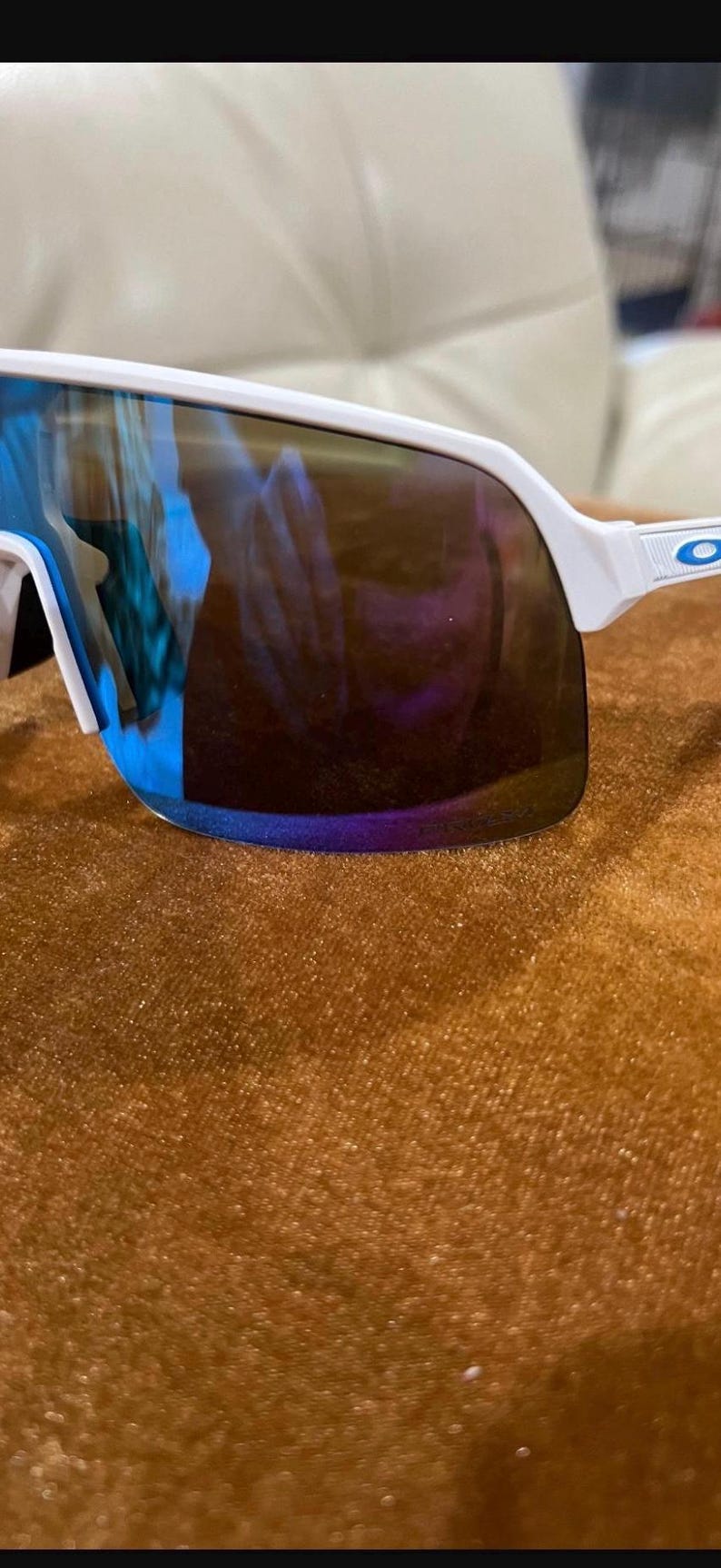 May include: White-framed sunglasses with a wraparound design. The lenses have a mirrored finish with blue and purple hues. The sunglasses are set against a brown textured surface.