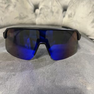 May include: Black sunglasses with a single-lens design and blue mirrored lenses. The sunglasses have a sleek, modern style, suitable for outdoor activities. The frame is a matte black color.