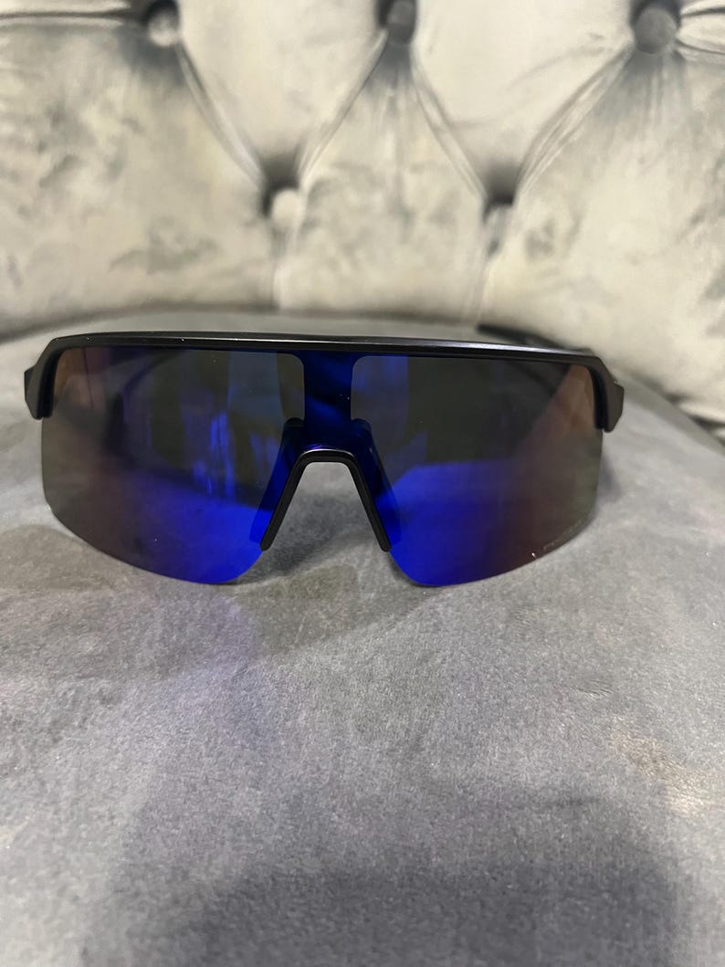 May include: Black sunglasses with a single-lens design and blue mirrored lenses. The sunglasses have a sleek, modern style, suitable for outdoor activities. The frame is a matte black color.