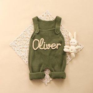 Puede incluir: Mono de bebé de punto verde oliva con el nombre "Oliver" en un color más claro. El mono tiene cierres de botón en las correas y en la entrepierna. Un pequeño conejito de peluche blanco está al lado del mono.