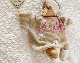 May include: A beige knit sweater featuring pink embroidered cursive text and white floral accents, worn with white socks and a white headband with a large bow. A wooden balloon-shaped sign with the text 2 months is positioned nearby.