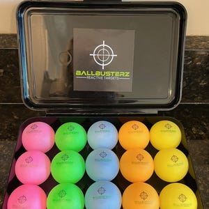 May include: A black case with a white target logo that says "Ballbusterz - Reactive Targets". The case is open and contains 18 colourful balls with a target printed on them. The balls are in a variety of colours, including pink, green, blue, yellow, and orange.
