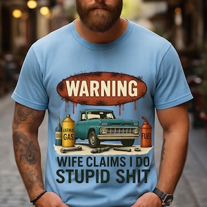 Puede incluir: Camiseta azul claro con un diseño gráfico de una camioneta turquesa, bidones de aceite y combustible, y herramientas. El texto dice "WARNING" y "WIFE CLAIMS I DO STUPID SHIT".