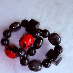 May include: A beaded necklace featuring a mix of black and red beads. The beads vary in shape, including round, oval, and rectangular. The red beads have black speckles. The necklace is displayed on a white marble surface.