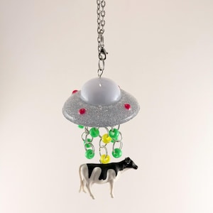 May include: A silver glitter UFO pendant with red dots and a white dome is suspended from a chain. Green and yellow beads hang below, 'beaming up' a black and white cow figurine. A whimsical, novelty item.