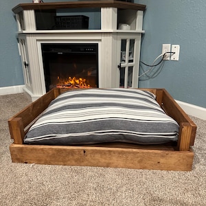 May include: A wooden dog bed with a striped gray and white cushion. The bed is made of natural wood and has a rustic finish.