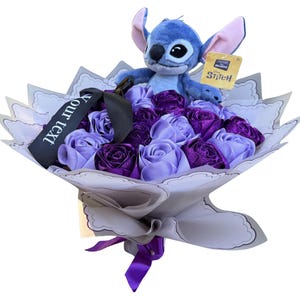 Op de afbeelding: Een boeket van paarse en lavendelkleurige satijnen rozen met een pluche Stitch-speelgoed erop. Het boeket is verpakt in wit en grijs papier, vastgebonden met een paars lint en bevat een zwart lint met de tekst "Your text".