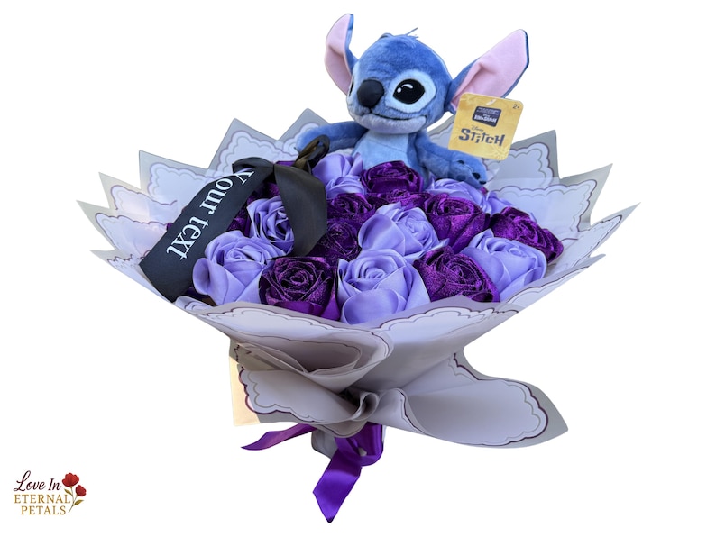 Op de afbeelding: Een boeket van paarse en lavendelkleurige satijnen rozen met een pluche Stitch-speelgoed erop. Het boeket is verpakt in wit en grijs papier, vastgebonden met een paars lint en bevat een zwart lint met de tekst "Your text".