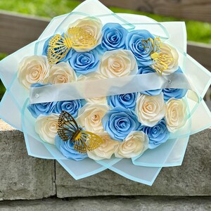 May include: A bouquet of paper roses in shades of blue and cream, accented with gold butterfly cutouts. The flowers are arranged in a circular shape and wrapped in light blue tissue paper, tied with a white ribbon.