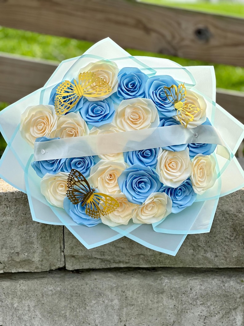 May include: A bouquet of paper roses in shades of blue and cream, accented with gold butterfly cutouts. The flowers are arranged in a circular shape and wrapped in light blue tissue paper, tied with a white ribbon.