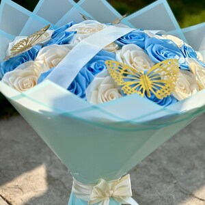 May include: A bouquet of blue and cream-colored roses, wrapped in light blue paper and adorned with gold butterfly accents. A cream-colored ribbon and bow secure the base of the bouquet, creating an elegant floral arrangement.