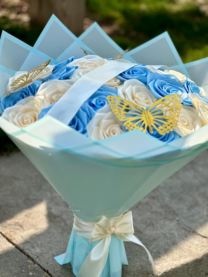 May include: A bouquet of blue and cream-colored roses, wrapped in light blue paper and adorned with gold butterfly accents. A cream-colored ribbon and bow secure the base of the bouquet, creating an elegant floral arrangement.