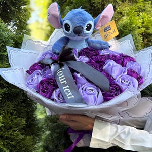 Op de afbeelding: Een boeket van paarse en lavendelkleurige stoffen rozen, bekroond met een blauw Stitch-knuffel. Het boeket is verpakt in wit papier met een decoratieve rand en een zwart lint. Een label met "Stitch" is bevestigd.