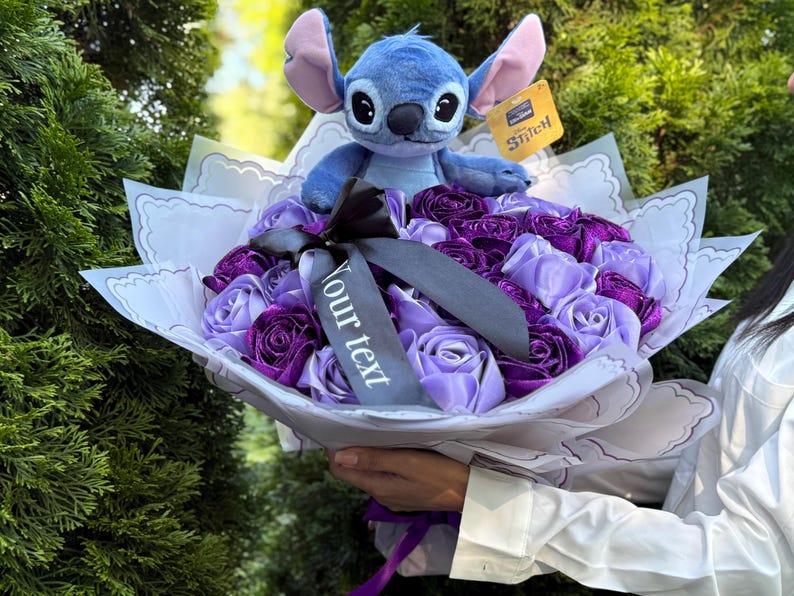 Op de afbeelding: Een boeket van paarse en lavendelkleurige stoffen rozen, bekroond met een blauw Stitch-knuffel. Het boeket is verpakt in wit papier met een decoratieve rand en een zwart lint. Een label met "Stitch" is bevestigd.