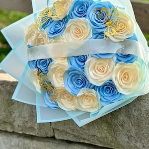 May include: A bouquet of paper roses in shades of light blue and cream, accented with gold butterfly embellishments. The flowers are wrapped in light blue and white tissue paper, tied with a white ribbon.