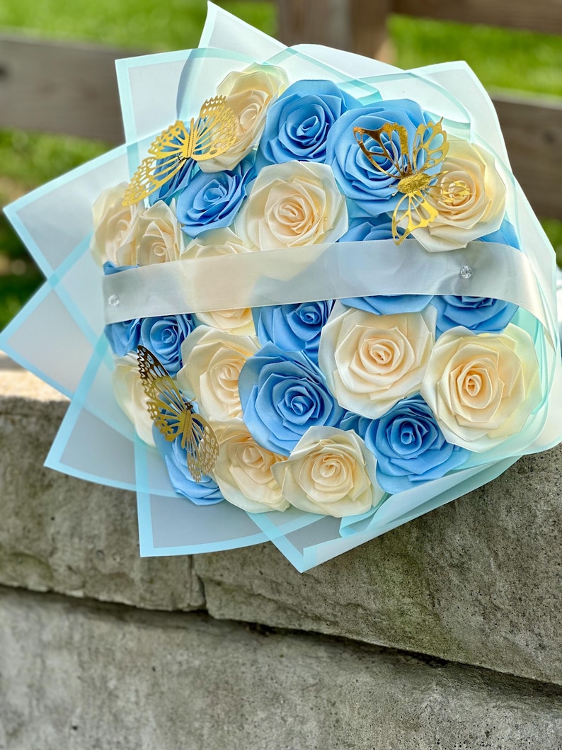 May include: A bouquet of paper roses in shades of light blue and cream, accented with gold butterfly embellishments. The flowers are wrapped in light blue and white tissue paper, tied with a white ribbon.