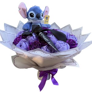 Op de afbeelding: Een boeket met een Stitch-knuffel bovenop paarse en lavendelkleurige stoffen rozen. Het boeket is verpakt in decoratief papier en vastgebonden met een paars lint. Een zwart lint met de tekst "Your Text" is ook inbegrepen.