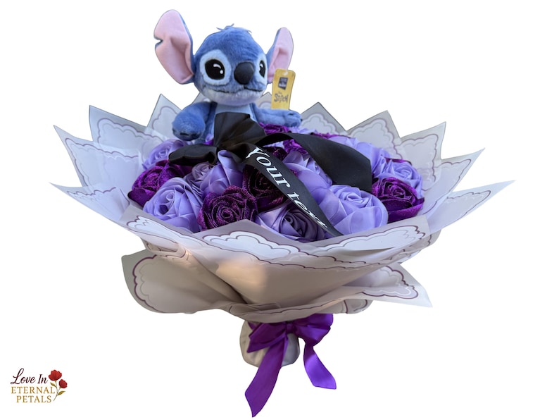 Op de afbeelding: Een boeket met een Stitch-knuffel bovenop paarse en lavendelkleurige stoffen rozen. Het boeket is verpakt in decoratief papier en vastgebonden met een paars lint. Een zwart lint met de tekst "Your Text" is ook inbegrepen.