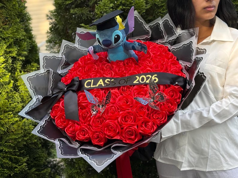 May include: A graduation bouquet featuring a plush Stitch toy wearing a graduation cap, set atop a bed of red roses. The bouquet includes a black ribbon with "CLASS OF 2026" and decorative butterflies. The bouquet is wrapped in black and white paper.