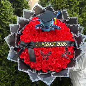 May include: A graduation bouquet featuring a plush Stitch toy wearing a graduation cap, surrounded by red roses. The bouquet includes a black ribbon with "CLASS OF 2026" and decorative butterflies. The arrangement is wrapped in black and white paper.