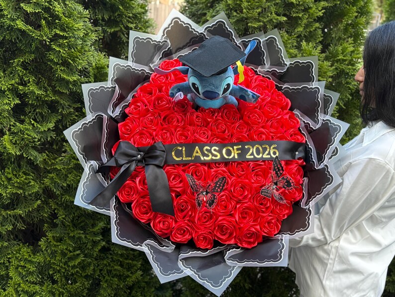 May include: A graduation bouquet featuring a plush Stitch toy wearing a graduation cap, surrounded by red roses. The bouquet includes a black ribbon with "CLASS OF 2026" and decorative butterflies. The arrangement is wrapped in black and white paper.