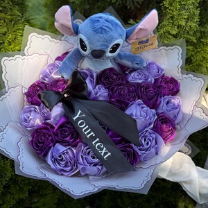 Op de afbeelding: Een boeket van paarse en lavendelkleurige rozen van lint met een blauw Stitch-knuffel. Het boeket is verpakt in wit papier en heeft een zwart lint met de tekst "Your text".