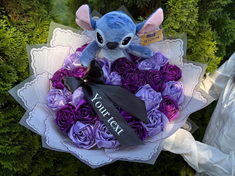 Op de afbeelding: Een boeket van paarse en lavendelkleurige rozen van lint met een blauw Stitch-knuffel. Het boeket is verpakt in wit papier en heeft een zwart lint met de tekst "Your text".