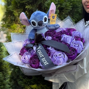Op de afbeelding: Een boeket van paarse en lavendelkleurige stoffen rozen, verpakt in wit papier met een geschulpte rand. Een blauw Stitch-knuffelbeest zit bovenop de bloemen en houdt een klein label vast. Een zwart lint met de tekst "Your text" is om het boeket gebonden.