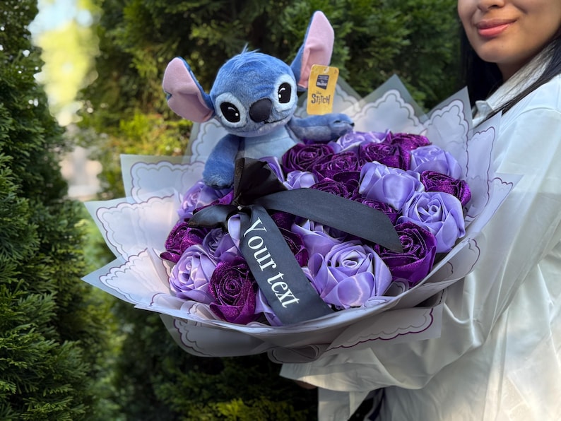 Op de afbeelding: Een boeket van paarse en lavendelkleurige stoffen rozen, verpakt in wit papier met een geschulpte rand. Een blauw Stitch-knuffelbeest zit bovenop de bloemen en houdt een klein label vast. Een zwart lint met de tekst "Your text" is om het boeket gebonden.