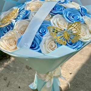 May include: A bouquet of artificial roses in shades of blue and cream, accented with gold butterfly decorations. The flowers are wrapped in light blue paper and tied with a cream ribbon, creating a visually appealing floral arrangement.