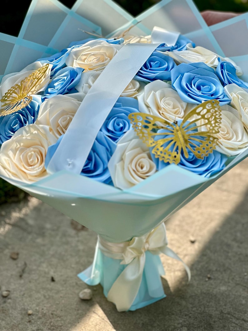May include: A bouquet of artificial roses in shades of blue and cream, accented with gold butterfly decorations. The flowers are wrapped in light blue paper and tied with a cream ribbon, creating a visually appealing floral arrangement.