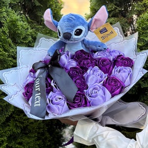 Op de afbeelding: Een boeket met paarse en lavendelkleurige rozen van lint, bekroond door een blauw Stitch-knuffel. Het boeket is verpakt in wit papier met decoratieve randen en een zwart lint met de tekst "our text". Een Stitch-label is bevestigd.