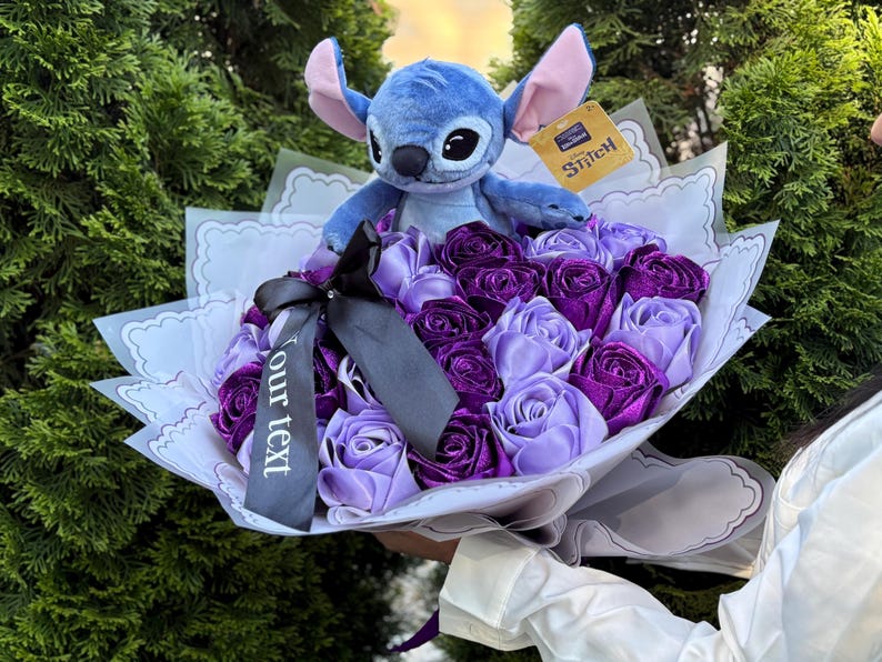 Op de afbeelding: Een boeket met paarse en lavendelkleurige rozen van lint, bekroond door een blauw Stitch-knuffel. Het boeket is verpakt in wit papier met decoratieve randen en een zwart lint met de tekst "our text". Een Stitch-label is bevestigd.