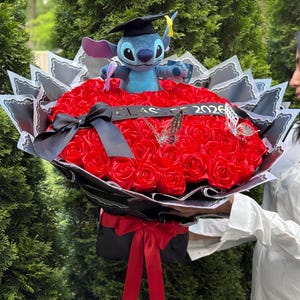 May include: A bouquet of red roses with a plush Stitch toy wearing a graduation cap. The bouquet is decorated with black and white paper, a black ribbon, and butterfly accents. The year 2026 is displayed on a ribbon.
