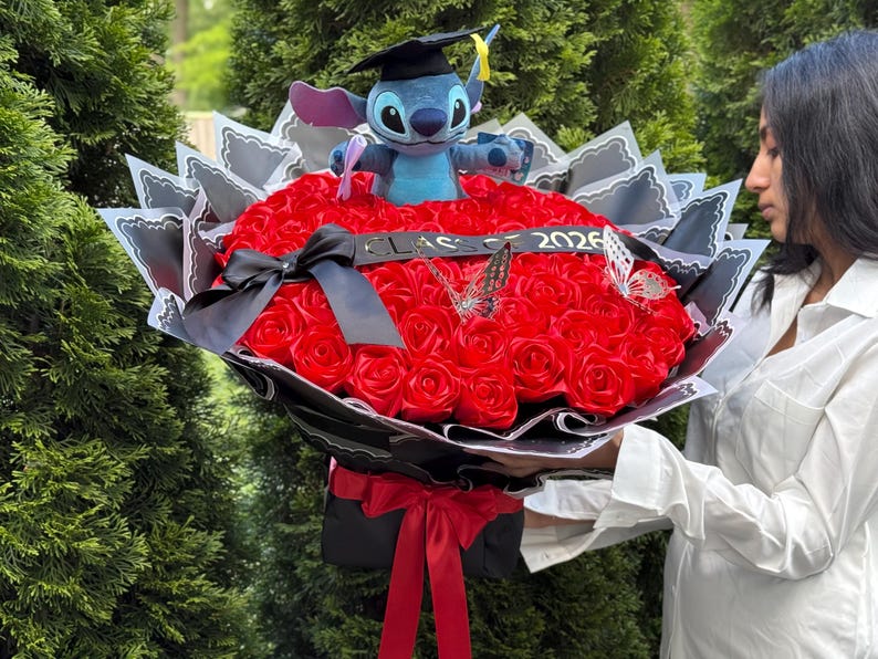 May include: A bouquet of red roses with a plush Stitch toy wearing a graduation cap. The bouquet is decorated with black and white paper, a black ribbon, and butterfly accents. The year 2026 is displayed on a ribbon.