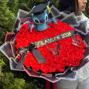 May include: A graduation bouquet featuring a plush Stitch toy wearing a graduation cap, surrounded by red roses. The bouquet includes a black ribbon with "CLASS OF 2026" and decorative butterflies. The arrangement is wrapped in black and white paper.