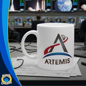 May include: White ceramic mug featuring the Artemis logo in black, red, and blue. The logo includes a stylized letter A with a sphere and a curved red line. The word "ARTEMIS" is printed below the logo in a dark blue font.