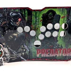 Puede incluir: Panel de control de arcade personalizado con el tema Predator. El panel tiene un fondo negro y verde con ilustraciones de Predator y la palabra "PREDATOR". Incluye botones plateados y un borde rojo. Es visible una pegatina de "Deep Sea Customs".