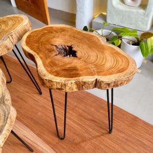 May include: A live edge side table featuring a natural wood slab top with a prominent central knot hole and visible grain patterns. The table is supported by three slender black metal hairpin legs, showcasing a raw and organic aesthetic.